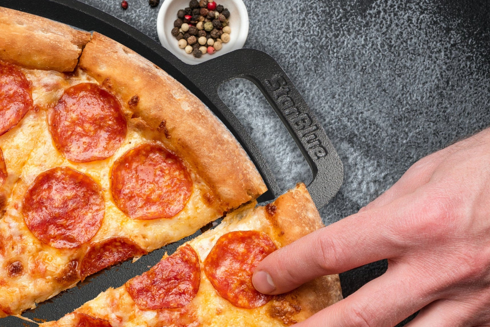 Pepperoni pizza on a pan with hands taking slices, surrounded by tomatoes and basil.